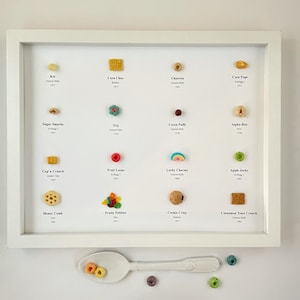 May include: A white frame with a collection of miniature cereal boxes. Each box is labeled with the cereal name and the company that makes it. The cereal boxes are arranged in rows of four. There is a white spoon with a few cereal pieces on it below the frame.