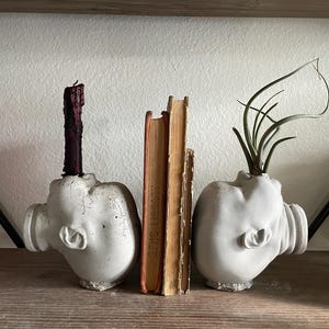 May include: Two white ceramic bookends shaped like human heads, each holding a book. The bookend on the left has a black candle in it, and the bookend on the right has a green air plant in it.
