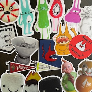 May include: A collection of colorful stickers featuring various cartoon characters, including a cat wearing a cone, a red devil, a marshmallow, and a black and white character with an X on its forehead. The stickers are on a wooden surface. The text on the stickers includes "Hungry", "Want Believe", "Aren't I Snazzy?" and "Employee of the Month".