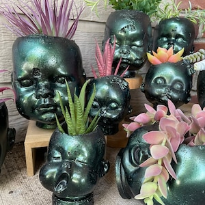 May include: A collection of doll head planters in a dark green color with various succulents and plants growing inside. The planters are made to look like vintage dolls with painted features.