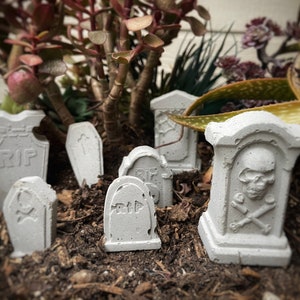 May include: A collection of miniature white concrete tombstones with the words "RIP" on them. Some tombstones have a skull and crossbones design. The tombstones are arranged in a small garden setting.