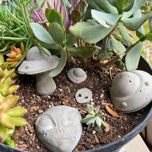 May include: A black planter pot filled with succulents and small concrete alien and mushroom figurines. The planter is sitting on a light brown surface.