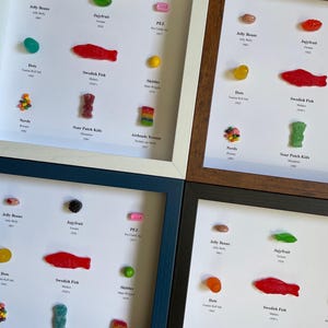 May include: Four white picture frames with black mats, each containing a collection of colorful candies. The candies are arranged in a grid pattern and labeled with their names and years of introduction. The candies include Jelly Beans, Jujyfruit, PEZ, Skittles, Dots, Swedish Fish, Nerds, Sour Patch Kids, and Airheads Xtreme.