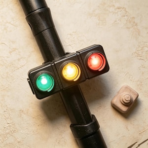 May include: A black cylindrical pole features a rectangular attachment with three circular lights arranged horizontally. The lights are illuminated in green, yellow, and red from left to right. A small, light-coloured, rectangular wooden block sits nearby.