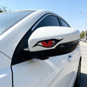 Peut inclure: Rétroviseur latéral de voiture blanc avec un autocollant en forme d'œil rouge et noir. L'autocollant est sur le boîtier du miroir blanc, avec un contour noir et un iris rouge. La voiture est garée dans la rue.