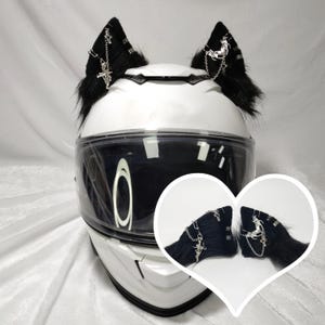 May include: White motorcycle helmet with black faux fur cat ears with silver chain and cross accents. The helmet has a clear visor.