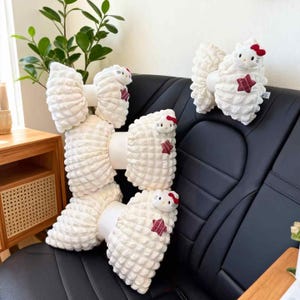 May include: White plush bow-shaped car seat cushions with a textured surface. Each bow features a small Hello Kitty character with a red bow and a red star accent. The cushions are arranged on a black car seat.