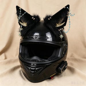 Może przedstawiać: Błyszczący czarny kask motocyklowy typu full-face z dwoma dużymi, stojącymi kocimi uszami z czarnego sztucznego futra. Uszy ozdobione są złotymi metalowymi zawieszkami w kształcie skrzydeł, łańcuszkami z jasnoniebieskimi kryształkami oraz gwiazdkami i księżycami.