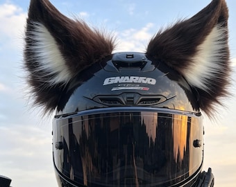 Może przedstawiać: Błyszczący czarny kask motocyklowy z dużymi, stojącymi uszami zwierzęcymi ze sztucznego futra na górze. Uszy są ciemnobrązowe z białym futerkiem wewnątrz. Kask posiada ciemną przyciemnianą szybkę oraz biały napis z nazwą marki GNARKO na przedniej części.