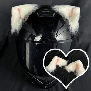 Peut inclure: Un casque de moto noir avec des oreilles de chat en fausse fourrure blanche fixées sur le dessus. Le casque a une visière transparente et une jugulaire noire. Les oreilles de chat sont douces et moelleuses.
