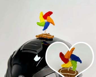 May include: A colorful, crocheted pinwheel atop a black motorcycle helmet. The pinwheel features red, orange, yellow, green, and blue blades. The base is brown and the pinwheel is attached to a wooden stick.