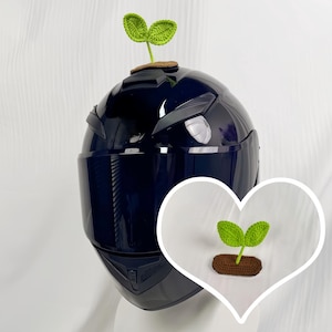 Peut inclure: Un casque de moto bleu foncé avec une pousse verte crochetée attachée au sommet. La pousse a deux feuilles vertes et une base marron. Une version plus petite de la pousse est dans un graphique en forme de cœur blanc.