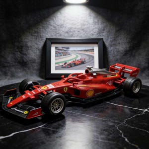 May include: A red Formula 1 race car model with black tyres and silver rims. The car has sponsor logos and a black cockpit. A framed picture of a race car on a track is in the background.