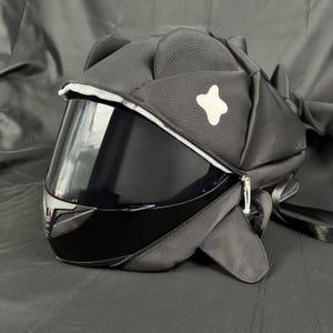May include: A black motorcycle helmet with a tinted visor, enclosed in a custom black fabric cover. The cover has a white star-shaped patch and a zip closure. The helmet has a sleek, modern design.