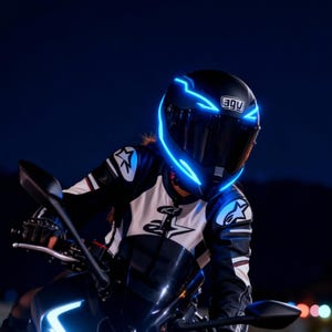May include: Black motorcycle helmet with bright blue neon accents. The helmet has the brand name "AGV" on the front. The rider is wearing a black and white motorcycle jacket. The image is taken at night.