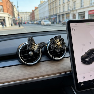 May include: Two black car air vent decorations, one shaped like a butterfly and the other a flower. The butterfly has gold accents, and the flower has gold detailing in the center. They are set against a car dashboard with a digital display.
