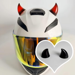 May include: White motorcycle helmet with black detailing and a gold visor. The helmet features red and black devil horns. A separate image shows a pair of black horns.