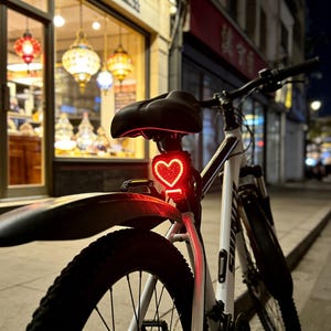 Op de afbeelding: Een witte fiets met een zwart zadel en een rood hartvormig achterlicht. Het licht is aan en de fiets staat 's nachts op een stoep. De achtergrond omvat een winkel met decoratieve lichten.