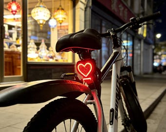 May include: A white bicycle with a black seat and a red heart-shaped taillight. The taillight is illuminated, and the bicycle is parked on a sidewalk at night. The background includes a storefront with decorative lights.