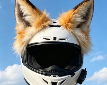 Może przedstawiać: Biały kask motocyklowy typu full-face z dużymi, pluszowymi uszami lisa ze sztucznego futra na górze. Uszy mają mieszankę pomarańczowego, kremowego i ciemnobrązowego futra. Kask posiada czarny wizjer, otwory wentylacyjne oraz małe czarne urządzenie komunikacyjne.