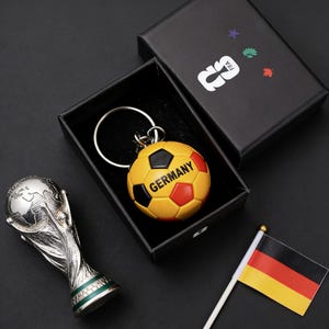 Puede incluir: Un llavero de balón de fútbol amarillo, negro y rojo con la inscripción "GERMANY", presentado en una caja negra. Un trofeo plateado y una pequeña bandera alemana también son visibles sobre una superficie negra.
