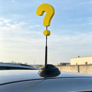 Puede incluir: Un adorno de antena de coche con forma de signo de interrogación amarillo. El adorno está montado sobre una base y un tallo negros, sobre un cielo azul. El techo del coche es visible en primer plano.