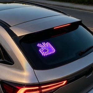 Puede incluir: Un coche hatchback plateado presenta una pantalla LED de color morado brillante en la ventana trasera. La pantalla muestra un gesto con la mano en el que el pulgar y el índice forman un círculo mientras los otros tres dedos permanecen verticales.