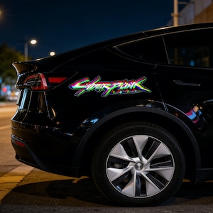 May include: Black car with a "Cyberpunk 2077" decal in neon green, blue, and pink. The decal is on the rear panel of the vehicle, near the window. The car is parked on a street at night.