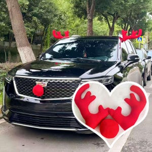 May include: A black SUV decorated with red reindeer antlers on the roof and a red nose on the front grille. The car is parked on a tree-lined street, suggesting a festive holiday theme. The image includes a heart-shaped inset with the antlers and nose.