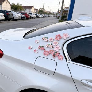 Puede incluir: Un coche blanco adornado con calcomanías de flores rosas y blancas y pétalos sueltos. Las calcomanías están dispuestas en el panel trasero, formando un diseño floral. El coche está estacionado en una calle con otros vehículos.