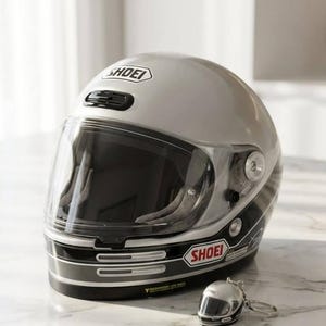 Könnte beinhalten: Silberner Motorradhelm mit klarem Visier und dem Markennamen "SHOEI". Ein passender Miniatur-Schlüsselanhänger-Helm ist ebenfalls vorhanden. Der Helm hat schwarz-weiße Akzente.