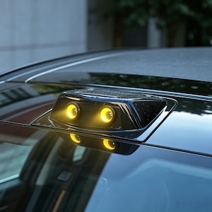 May include: A black automotive sensor with two illuminated yellow lights, mounted on the roof of a car. The sensor is rectangular with a slightly angled top, reflecting on the car's windshield.