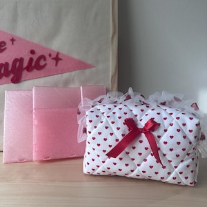 May include: A quilted white pouch featuring a repeating pattern of small red hearts, adorned with a dark red ribbon bow and a white ruffled lace trim. Beside it are two translucent pink rectangular plastic panels.
