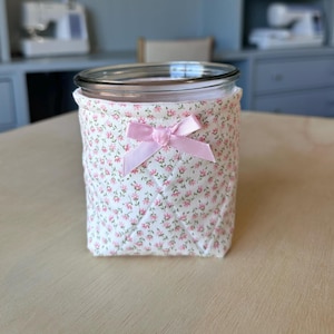 Może przedstawiać: Przezroczysty szklany słoik z pikowaną osłoną z tkaniny. Osłona jest biała z różowym kwiatowym wzorem i różową satynową kokardą. Słoik stoi na jasnobrązowej powierzchni.