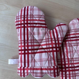 Puede incluir: Un par de guantes para horno de cuadros rojos y blancos con bordes festoneados. Los guantes están acolchados y tienen un lazo para colgar.