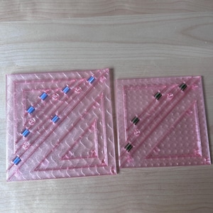 May include: Two pink quilting rulers with diagonal and square markings. The larger ruler has blue accents, while the smaller one has green accents. Both are made of clear plastic and are designed for precise fabric cutting and measuring.