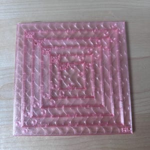 May include: A transparent pink square quilting template with a series of concentric squares and decorative patterns. The template is made of a clear, flexible material and is designed for quilting projects. The design includes curved lines and small circles.