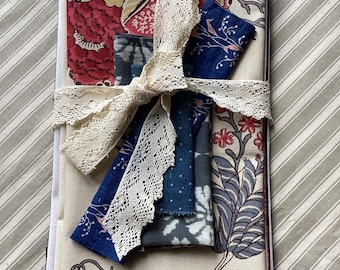 Könnte beinhalten: Ein stoffbezogenes Notizbuch, gebunden mit einem cremefarbenen Spitzenband. Das Notizbuch zeigt verschiedene gemusterte Stoffe in Rot, Blau und Creme mit floralen und abstrakten Designs. Mehrere Stoffmuster sind unter dem Band versteckt.