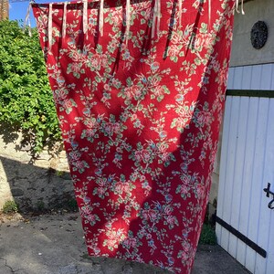 Könnte beinhalten: Ein großes, rotes Stoffpaneel mit einem sich wiederholenden Blumenmuster in Rosa, Grün und Blau. Der Stoff hängt an einer Kette mit weißen Bändern. Die Aufnahme wurde im Freien gemacht, mit einer Steinmauer und einer weißen Tür im Hintergrund.
