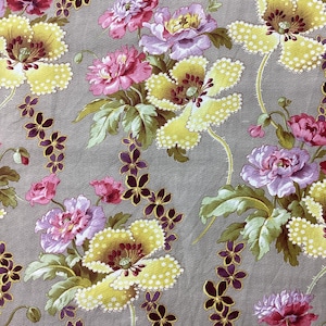 Op de afbeelding: Stof met een bloemmotief van grote gele en roze bloemen, paarse accenten en groene bladeren op een grijze achtergrond. De bloemen hebben een gestippelde textuur en het ontwerp bevat kleinere paarse bloemclusters.