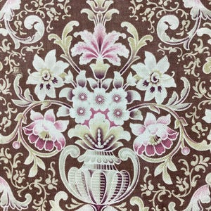 Könnte beinhalten: Nahaufnahme eines braunen Stoffes mit einem sich wiederholenden Blumenmuster. Das Design zeigt weiße, rosa und hellgrüne Blumen und Blätter, die in einem symmetrischen, verzierten Stil angeordnet sind. Der Hintergrund ist dunkelbraun.