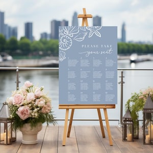 May include: A light blue seating chart with white seashell illustrations and the text "PLEASE TAKE your seat". The chart is displayed on a wooden easel, with floral arrangements and lanterns in the foreground.