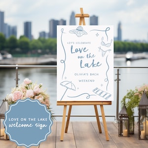 May include: A white welcome sign on a wooden easel with the text "Love on the Lake" and beach-themed illustrations in blue. The sign is for Olivia's Bach Weekend. The background includes a city skyline and water.