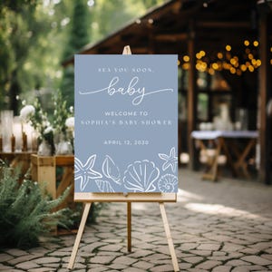 May include: A light blue baby shower welcome sign on a wooden easel. The sign reads "Sea You Soon, Baby" and welcomes guests to Sophia's baby shower on April 12, 2030. White seashell illustrations decorate the bottom.