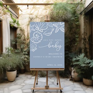 May include: A baby shower welcome sign on an easel. The sign is light blue with white seashell illustrations. Text reads "Sea you soon, baby. Welcome to Sophia's Baby Shower, April 12, 2030."
