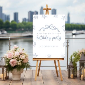 May include: A white sign on a wooden easel, with the text "WELCOME TO OLIVIA'S BIRTHDAY PARTY" in blue script. The sign is decorated with seashell and starfish illustrations. The date "SATURDAY, APRIL 12" is also displayed.