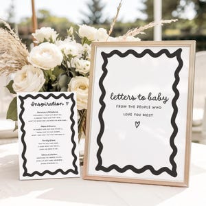 May include: Two framed signs with black wavy borders. One sign reads "Inspiration" and the other reads "letters to baby." Both signs have white backgrounds and black text. A bouquet of white roses and pampas grass is in the background.