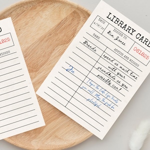 May include: Two vintage-style library cards on a wooden tray. The cards have a cream background with black lines and text, including the words "LIBRARY CARD" and "ADVICE AND WISHES." One card has handwritten notes in blue ink.