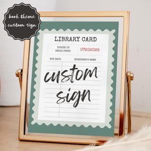 May include: A framed sign with a library card design. The sign features the words "custom sign" in a handwritten font. The library card includes the text "Issued to Emily Jones" and the date "07202025". The frame is gold.