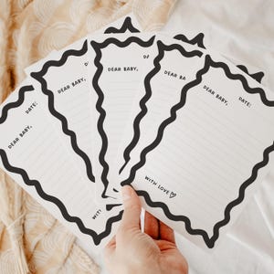 May include: A collection of white cards with black, wavy edges and lined interiors. The cards feature the text "Dear Baby," and "Date:" in black. One card has "With Love ♡" printed on it. The cards are held by a hand.